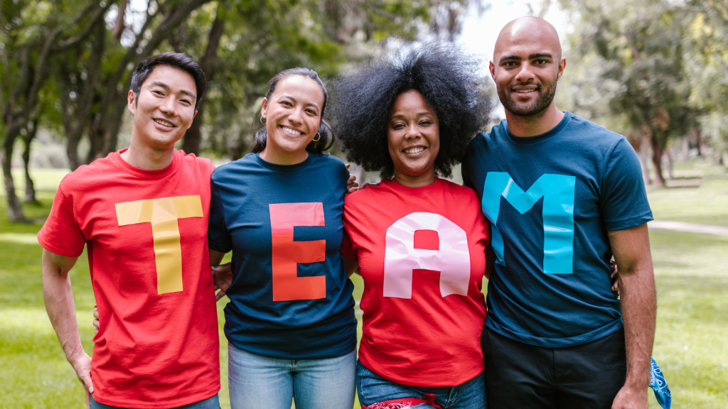 This is a photo of 4 people standing in a line. Each person has a t shirt on with a letter on it spelling out the word TEAM.